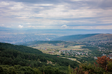 Naklejka premium Summer mountain view of Tbilisi, Georgia. Soft clouds cast gentle light over the city and its green landscape, with lush trees in the foreground, creating a calm, scenic atmosphere