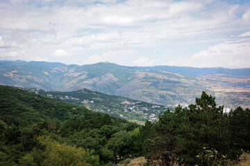 Naklejka premium Summer mountain view of Tbilisi, Georgia. Soft clouds cast gentle light over the city and its green landscape, with lush trees in the foreground, creating a calm, scenic atmosphere