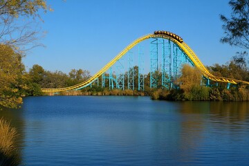 Fototapeta premium A vibrant roller coaster soaring over a tranquil lake, surrounded by trees under a clear blue sky, capturing the thrill of amusement park fun.