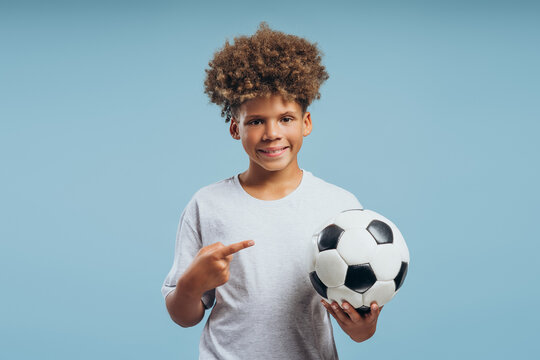 Cheerful, handsome African American boy with curly hair holding soccer ball, pointing finger at it
