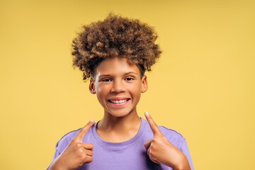 Attractive, smiling African American boy with curly hair pointing finger on teeth, looking at camera