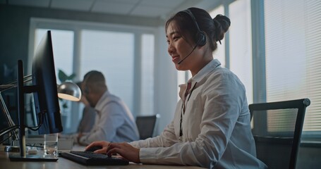 Call center office: Side view of friendly Asian female hotline operator talking on headset, using...