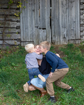 Portrait of a mother sitting on the grass in front of a wooden building with her two sons hugging and kissing her, Beckwith Township at Carleton Place in the Ottawa Valley; Ontario, Canada