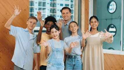 Smart teacher and diverse student standing while waving hand to camera. Skilled instructor with highschool children with mixed races in casual outfit greeting and smiling with cheerful. Edification.