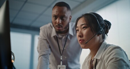 Modern call center office: Asian technical support operator in headset uses computer, talks to African American colleague. Two diverse helpdesk specialists working in online customer support service.