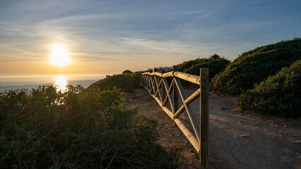Scenic view of the ocean and Benagil Beach at sunset along the coast of Portugal, with a wooden rail fence along a sandy trail at a lookout point; Lagoa, Faro, Portugal
