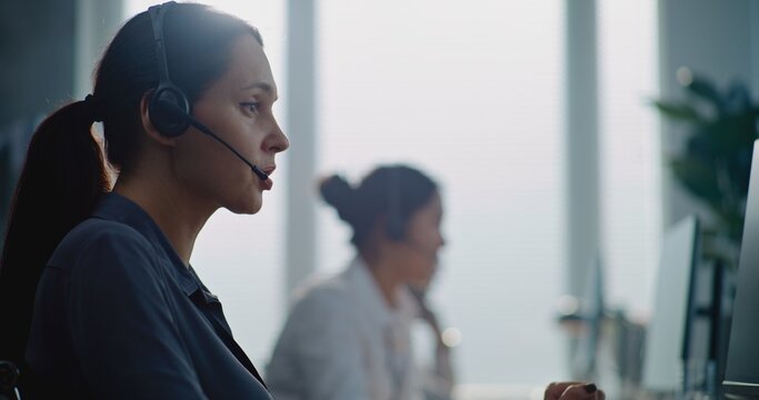 Call center office: Portrait of Caucasian female technical customer support operator working on computer, talking on headset, providing online help for client. Team of helpdesk specialists working.