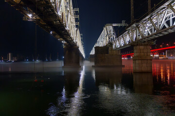 Steel bridge illuminated at night over calm river with reflections and mist