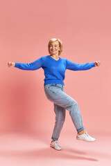 Full-length. Smiling, cheerful mature, elderly woman in jeans and sweater in motion, dancing against pink studio background. Concept of senior lifestyle, emotions, health, care, wellness