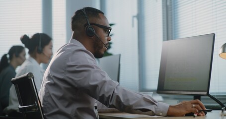 Obraz premium Modern call center office: Portrait of African American technical customer support specialist talking on headset, working on computer, solving problem, providing online help. Team of hotline operators