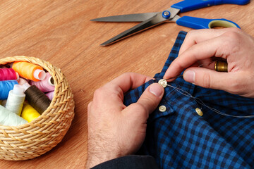 A person sewing a button on a blue plaid shirt with sewing tools and threads on a wooden table