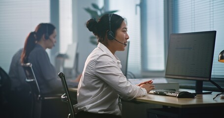 Multiethnic team of technical support operators working in modern call center office. Asian female helpdesk specialist in headset consults client via call, uses computer, providing customer service.