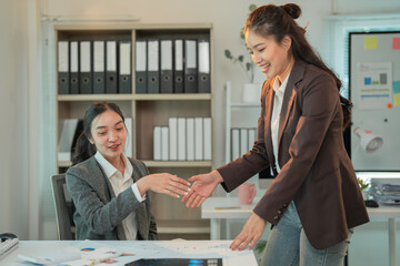Two asian businesswomen shaking hands over a desk with documents and charts, celebrating successful negotiation and partnership in a modern office environment