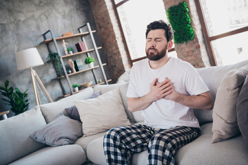 Photo of attractive man wear pajama sit on sofa arm touch chest heart attack pain hurt living room apartment indoors