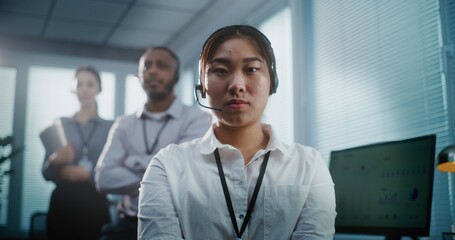 Obraz premium Modern call center control room: Close up portrait of serious Asian female hotline operator in headset looking at camera. Team of technical customer support specialists standing in the background.
