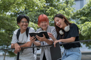 Three cheerful students are leaning on a railing, sharing a book and laughing together, enjoying...