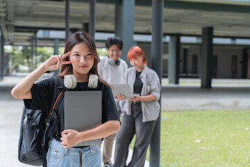 Young asian female student gesturing peace sign while holding a laptop with her friends in the...
