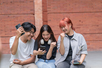 Three cheerful asian students are sitting together outdoors, expressing excitement and joy as they look at their smartphones, possibly celebrating academic achievement or good news