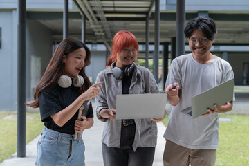 Three cheerful asian students are walking together outdoors, engaged in a lively discussion while using their laptops and sharing ideas on a university campus