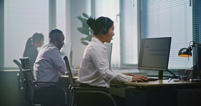 Multiethnic team of technical support operators working in modern call center office. Asian female helpdesk specialist talks on headset, uses computer, looks at camera providing customer service.