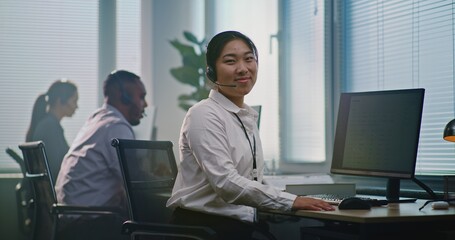Fototapeta premium Asian female technical support operator wearing headset, sits in front of computer, looks at camera providing customer service. Team of diverse helpdesk specialists work in modern call center office.
