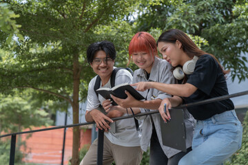 Three young asian students are leaning on a railing, sharing notes and studying together in a green urban park, enjoying the pleasant weather and collaborating on their education