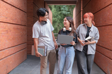 Three cheerful asian students are walking together in a university corridor, holding a laptop, books and folders, discussing their studies and enjoying their time on campus © Tj