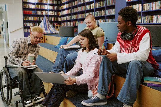 Group of students collaborating in modern library setting, with one student in wheelchair holding a coffee, others engaged with books and laptop