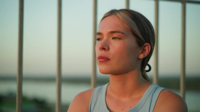 Close-up of young lady with thoughtful expression as sunlight reflects off her face, sitting near iron railing during sunset, creating a moody atmosphere with blurred background