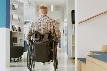 Man in wheelchair navigating through modern office hallway with backpack. White walls and carpeted stairs create an accessible environment