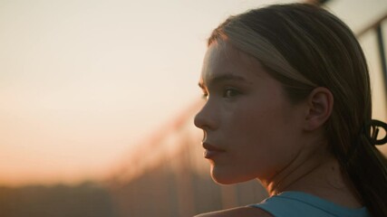 Close-up of young lady with moody expression looking back as sunlight reflects off her face, sitting near iron railing in the evening