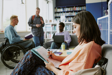 Group of students sitting and studying in a modern, bright library, engaging in conversation and using electronic devices. Multiracial individuals working collaboratively in inclusive setting