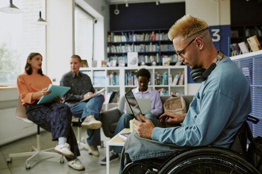 Group of diverse individuals engaging in study session in modern library workspace. Man in wheelchair focusing on work with laptop, while others discussing and taking notes