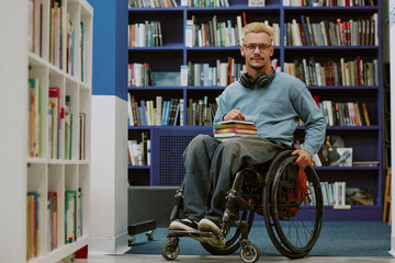 Young man sitting in wheelchair holding books while wearing headphones in library setting. He is...