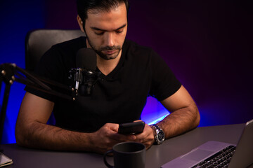 Broadcaster talking to listener on live streaming online, checking message from smartphone on social media in host channel by creative positive thinking concept at purple neon light studio. Surmise.