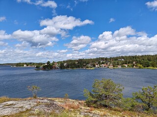 Scenic lakeside view outside of Stockholm under a bright blue sky with scattered clouds, surrounded by lush greenery and a distant shoreline