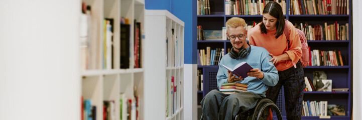 Engaging in a study session, two students explore a book in a library. One student is in a wheelchair while the other assists by looking over their shoulder with focus and interest