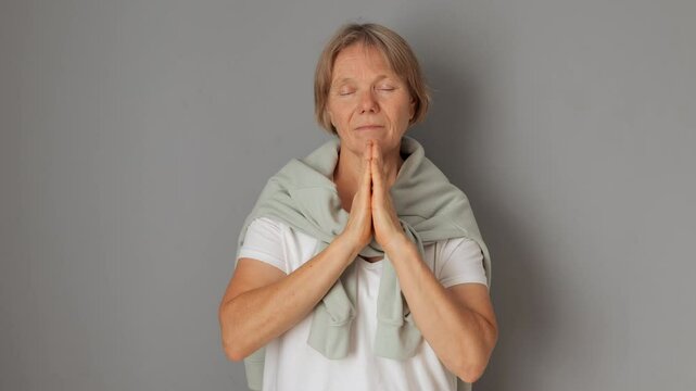 Senior woman with closed eyes and hands in prayer position expressing calm gratitude while pleading for hope and making heartfelt request isolated over gray background