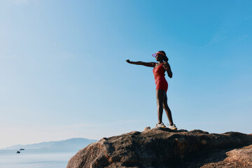 A person standing on a rock near the sea, wearing a red swimsuit and a hat, reaching out with their hand against a clear blue sky