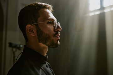 Medium close up of young handsome priest with round glasses in black robe looking straight