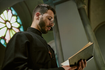 Fototapeta premium Low angle shot of young pastor with glasses and dressed in robe with wooden cross on chest reading Bible during service in local church