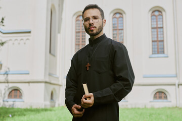 Portrait of young Caucasian priest dressed in black robe and wearing big wooden cross holding Bible...