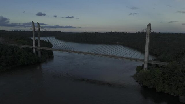 Drone flies to the left over Oiapoque River facing suspension bridge between Brazil and French Guyana at sunset
