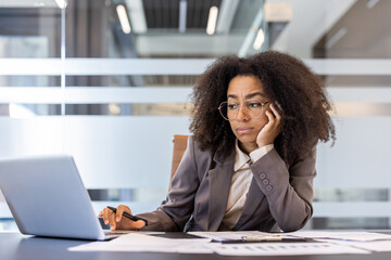 Tired young African American woman sitting in office at desk, holding head in hand and looking...