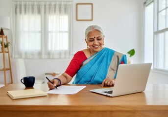 Smiling elderly Indian woman in traditional attire working on laptop at home office desk