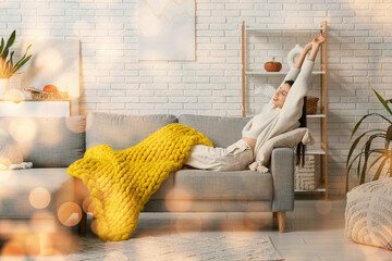 Beautiful young woman with warm yellow plaid stretching herself and lying on sofa in living room
