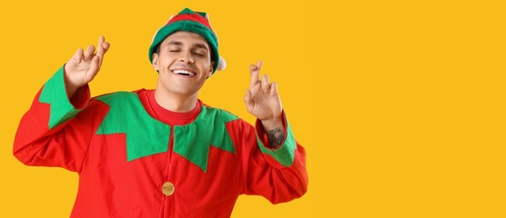 Young man in elf's costume and with crossed fingers on yellow background with space for text