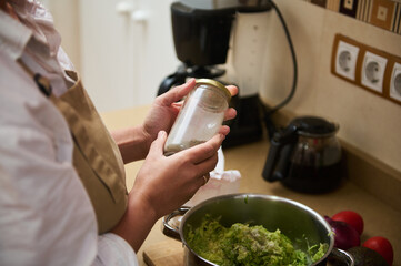 Person preparing homemade meal in cozy kitchen, wearing an apron and adding spices to a bowl of...
