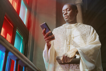 Low angle shot of African American priest in beautiful robe checking his smartphone while standing...
