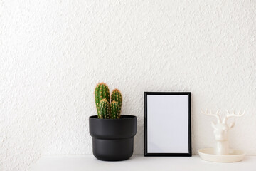 Beautiful blank photo frame, cactus in pot and deer figure on table near white wall